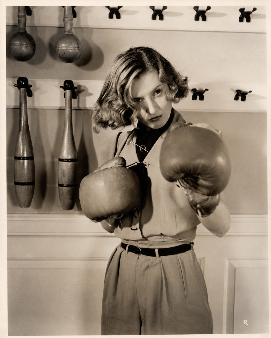 A woman in vintage attire wears boxing gloves and poses with one fist forward. Wooden clubs hang on the wall behind her in what appears to be an old-fashioned gym or training room.