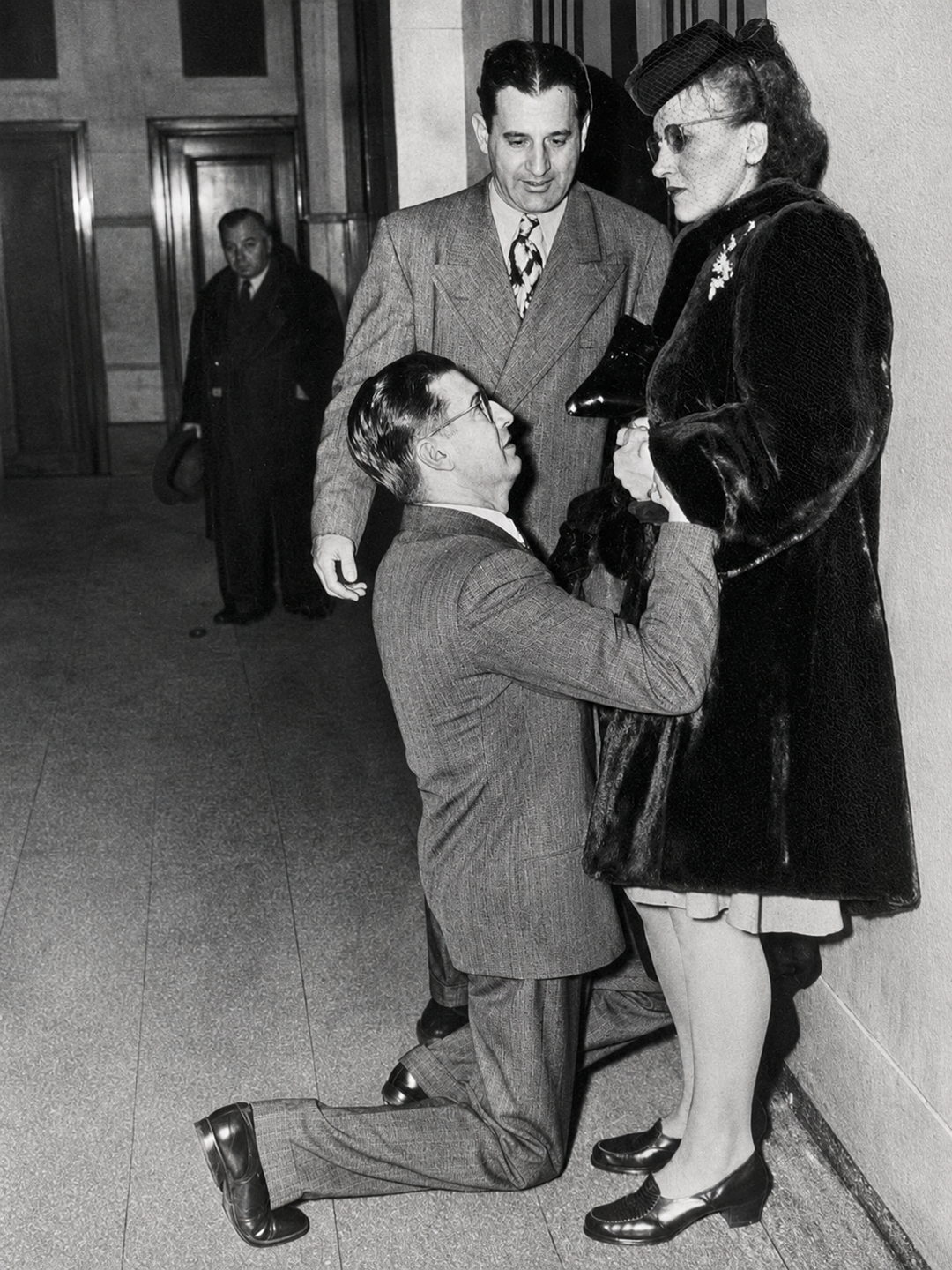 A man in a suit kneels on the floor, holding a woman's hands. The woman wears a fur coat and hat. Another man stands behind them. All three appear to be inside a building.