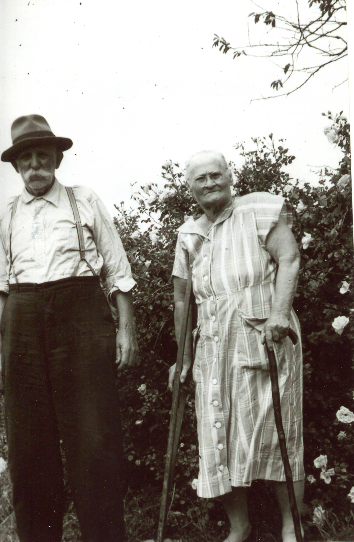 An elderly woman with a cane and an elderly man in a hat stand outdoors by bushes. Both wear plain clothes and look at the camera. The photo is black and white.