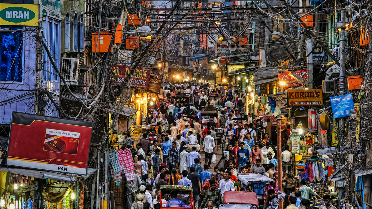 A crowded street market in an urban area, filled with people, shops, hanging signs, and tangled overhead wires. Bright lights from stalls illuminate the busy scene in the narrow, bustling alleyway.