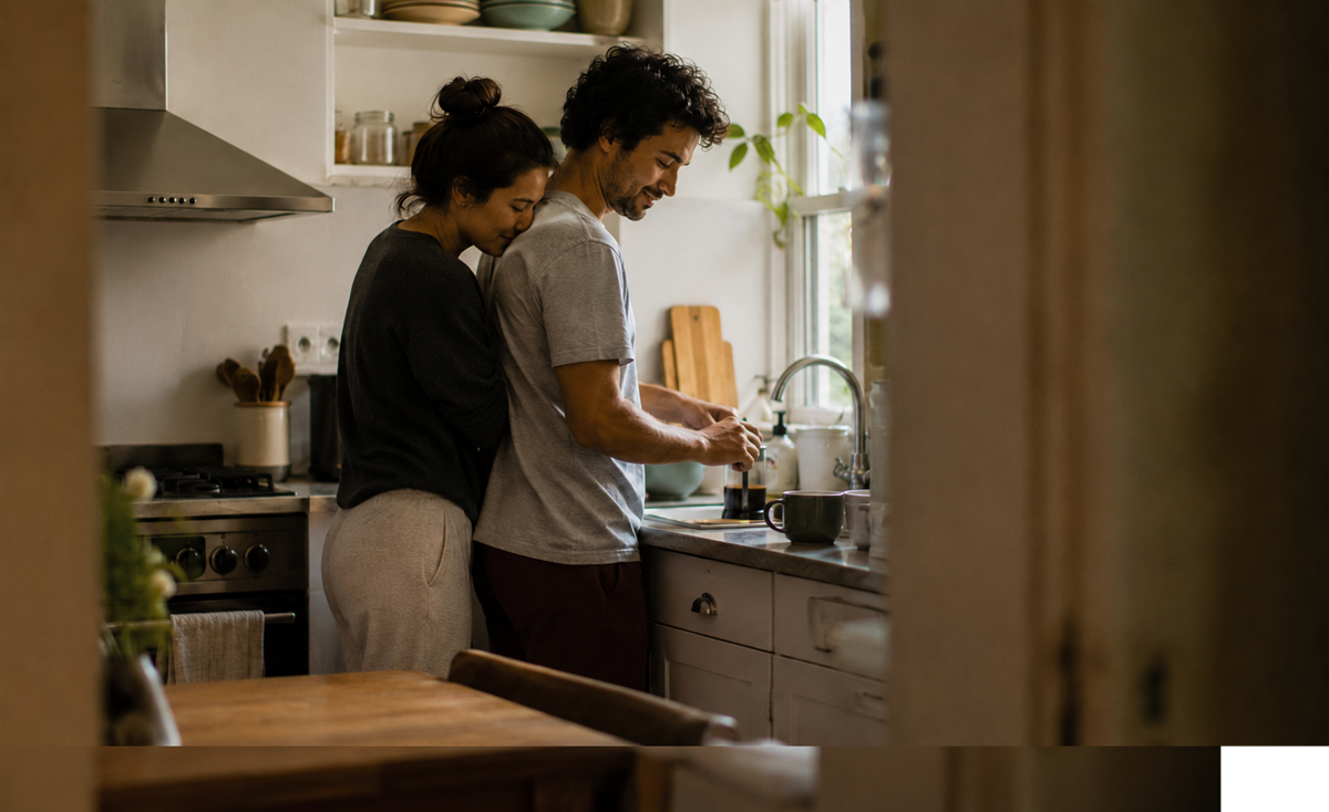 A couple stands together in a cozy kitchen. One person hugs the other from behind while they wash dishes at the sink. Warm light comes through the window, creating a relaxed, intimate atmosphere.