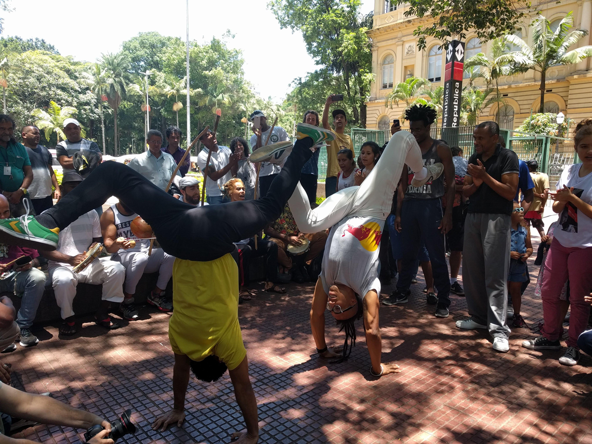 Two people perform capoeira acrobatics on a tiled plaza, surrounded by a crowd watching and clapping. The background features trees and a yellow building.