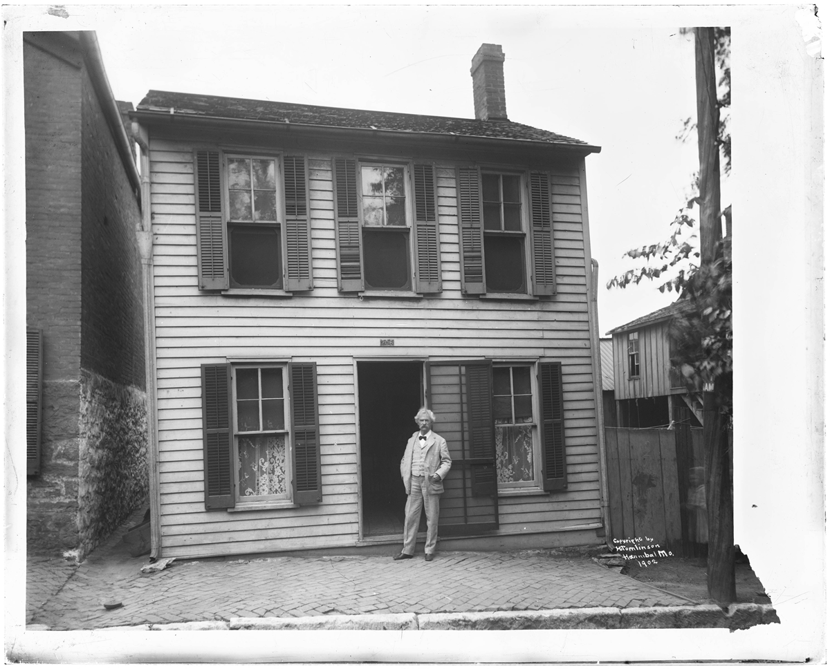 An older man with white hair and a mustache stands in the doorway of a two-story wooden house with shutters, on a brick sidewalk. The house has lace curtains in the windows.