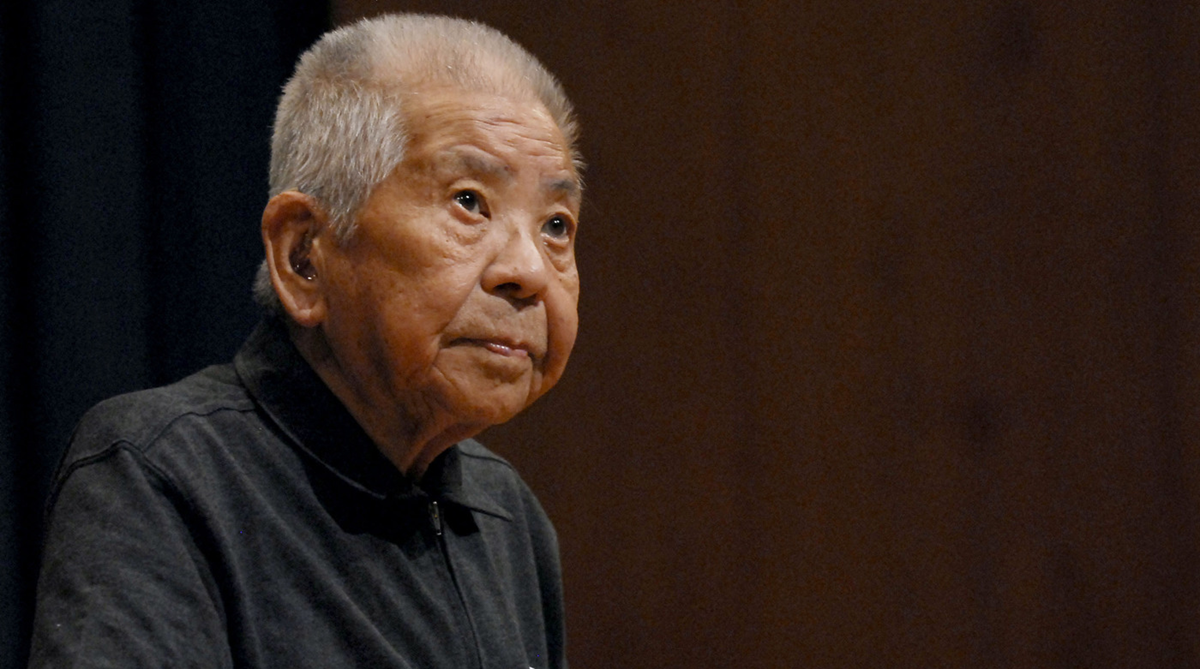 An elderly person with short white hair and a dark collared shirt looks slightly upward with a thoughtful expression, set against a plain dark background.
