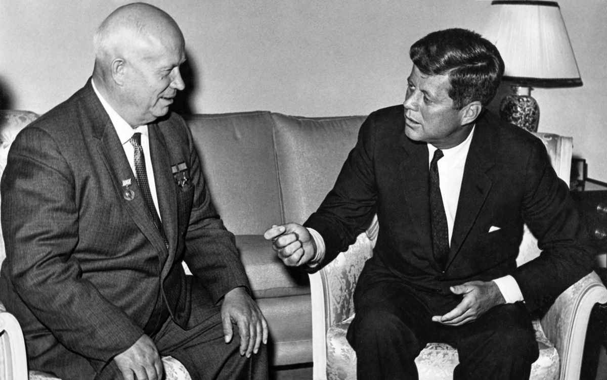 Two men in suits sit and talk on armchairs in a formal room. One man gestures with his hand while speaking; the other listens. A lamp and sofa are visible in the background.