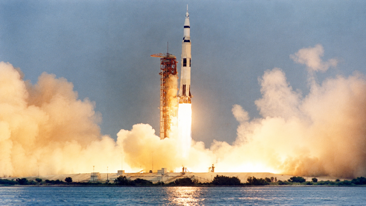 A large rocket launches from a platform, with bright flames and smoke billowing out, as it ascends into the sky near a body of water.
