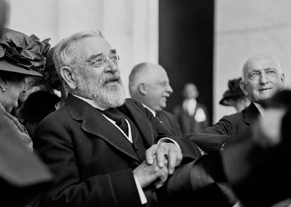 An older man with a white beard and glasses smiles while sitting among a group of people in formal attire, some wearing hats, during an outdoor event.