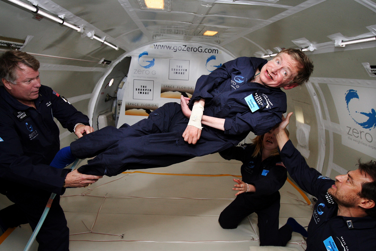 Four people float in a zero-gravity plane. One person in the center is being supported by three others, all wearing dark blue jumpsuits with patches. The interior is white, with "zero G" and logos visible on the walls.