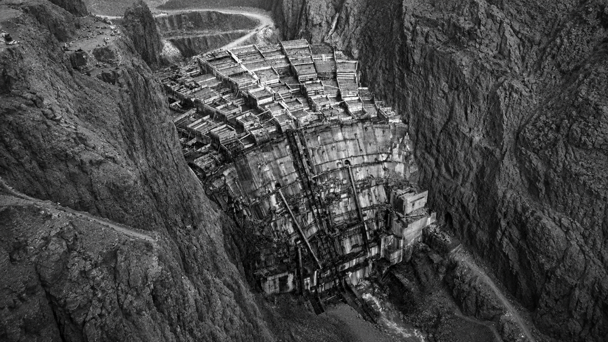 A large, partially constructed dam stands between steep rocky cliffs in a black and white photo, with construction materials and structures visible on the dam face.