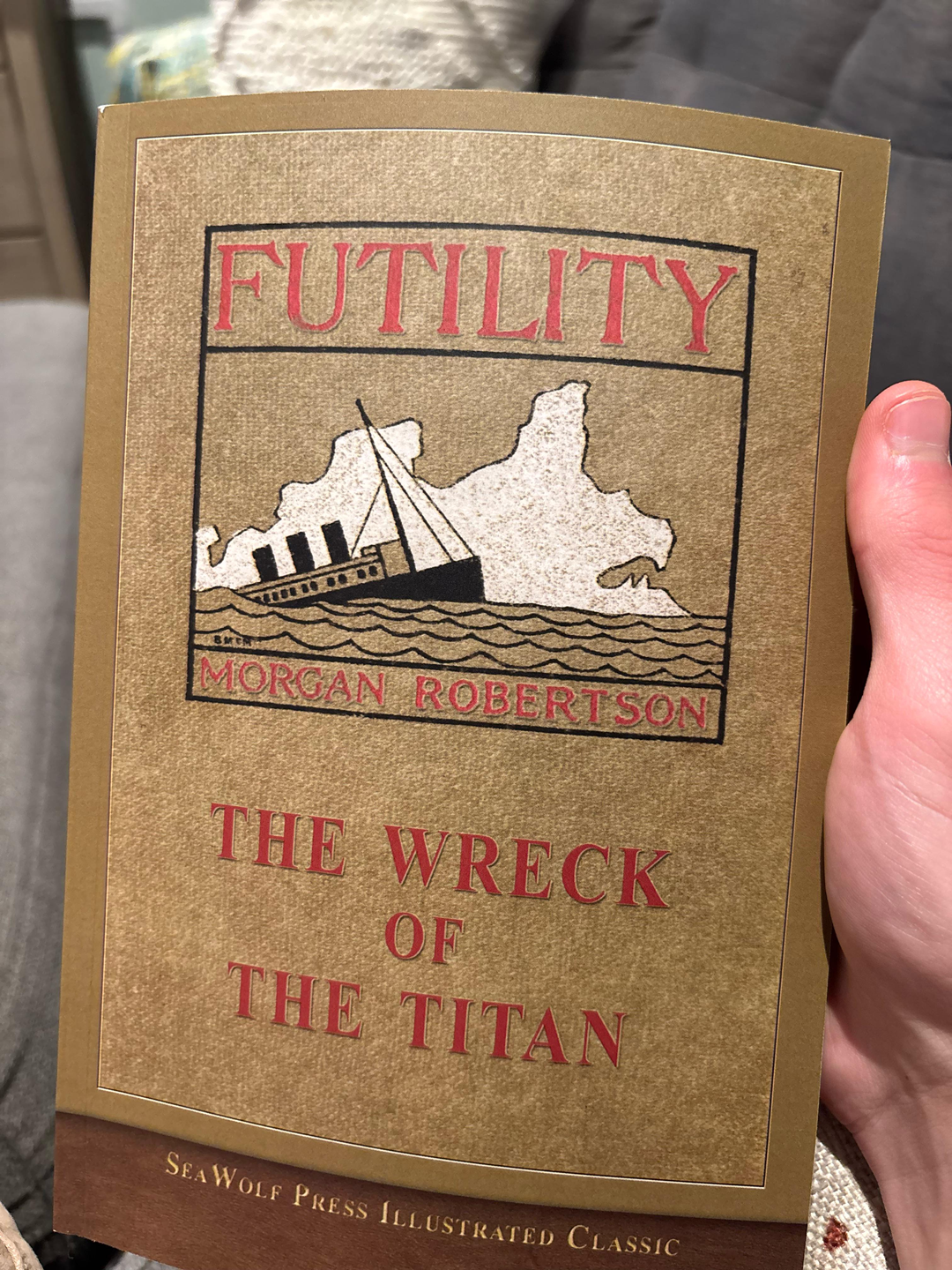 A hand holds a book titled "Futility: The Wreck of the Titan" by Morgan Robertson, featuring an illustration of a sinking ship on the cover.