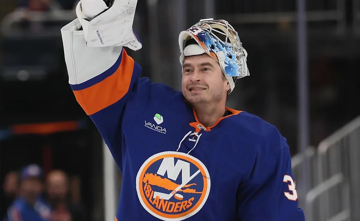 A hockey player in a blue New York Islanders jersey raises his gloved hand, with his helmet pushed up on his head, smiling on the ice.