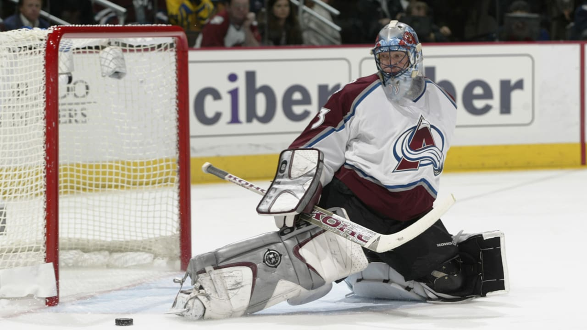 A Colorado Avalanche hockey goalie kneels on the ice in front of the net as a puck slides past him into the goal. The net is tilted and spectators are visible in the background.