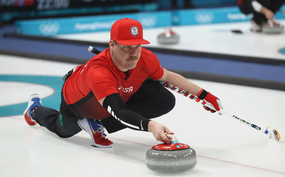 A curler in a red shirt and cap slides on the ice, holding a curling stone and broom, focused during a game.