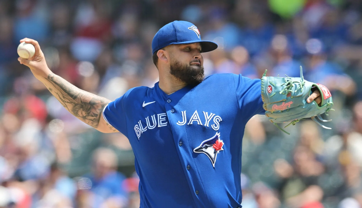 A baseball player in a blue Toronto Blue Jays uniform is pitching during a game, holding a baseball in his right hand and wearing a blue glove on his left.
