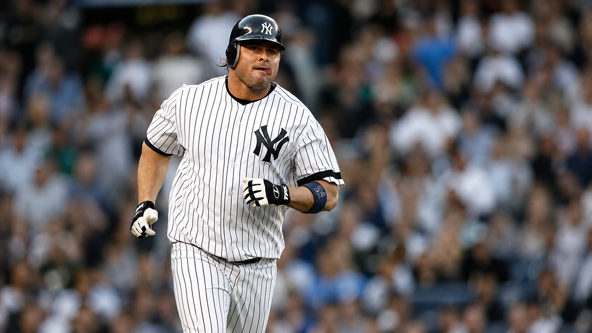 A New York Yankees baseball player in pinstripes runs on the field during a game, holding a bat, with a blurred crowd in the background.