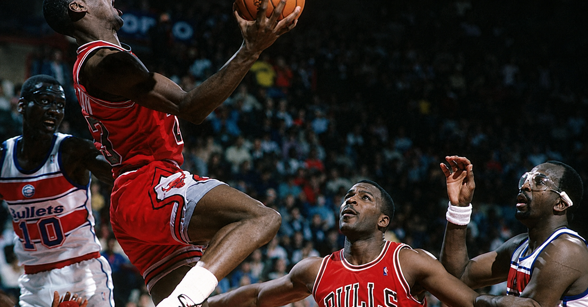 A basketball player in a red Chicago Bulls uniform leaps for a layup against Washington Bullets defenders during a game, with a teammate watching nearby.