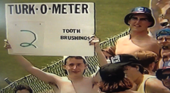 Two shirtless fans at a sports event hold a sign reading "TURK-O-METER 2 TOOTH BRUSHINGS." One wears a bucket hat, and there are more people and a chain-link fence in the background.