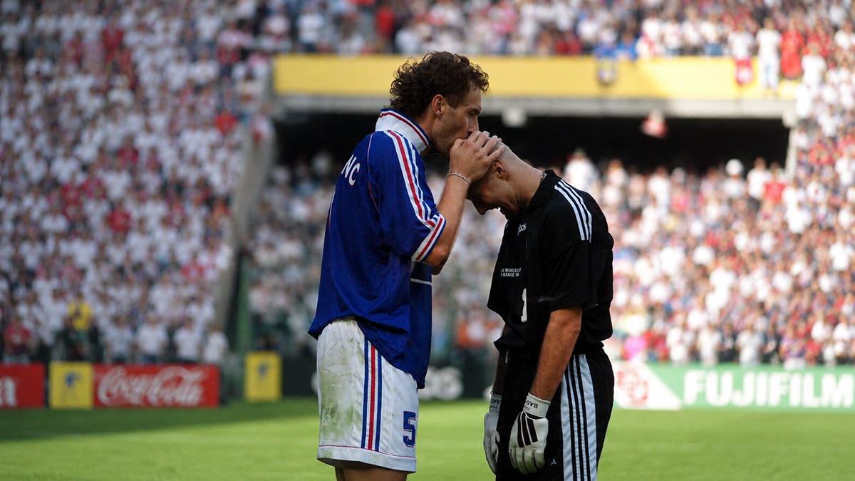 A soccer player in a blue jersey consoles a goalkeeper in black on the field, gently touching his forehead, with a blurred crowd in the background.