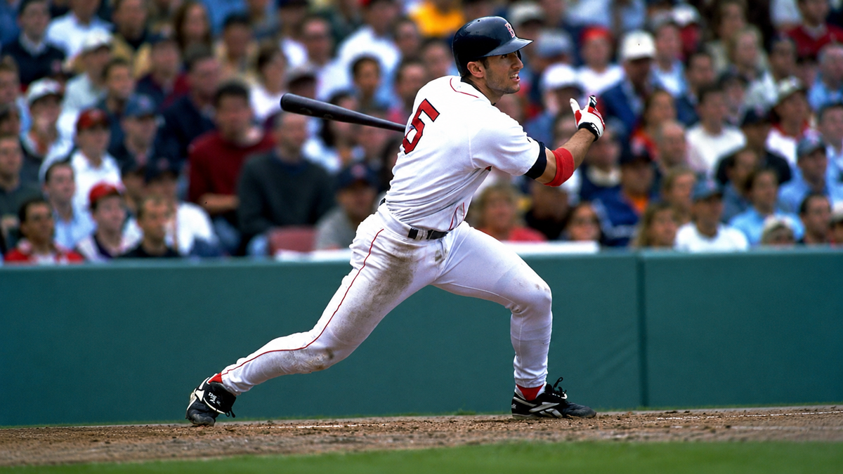 A baseball player in a white uniform swings a bat during a game, with a crowd of spectators watching from the stands behind him.