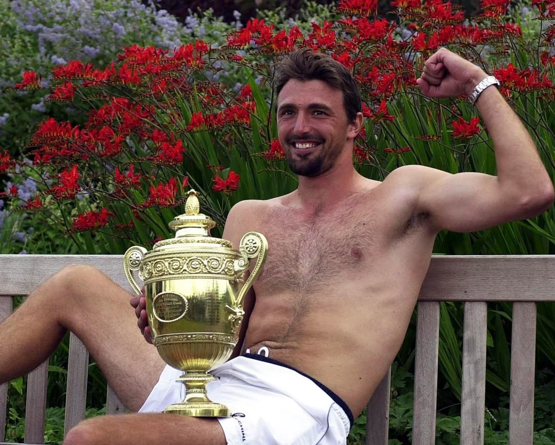 A smiling, shirtless man sits on a bench outdoors, flexing his right arm and holding a large gold trophy in his left hand. Bright red flowers and green plants fill the background.