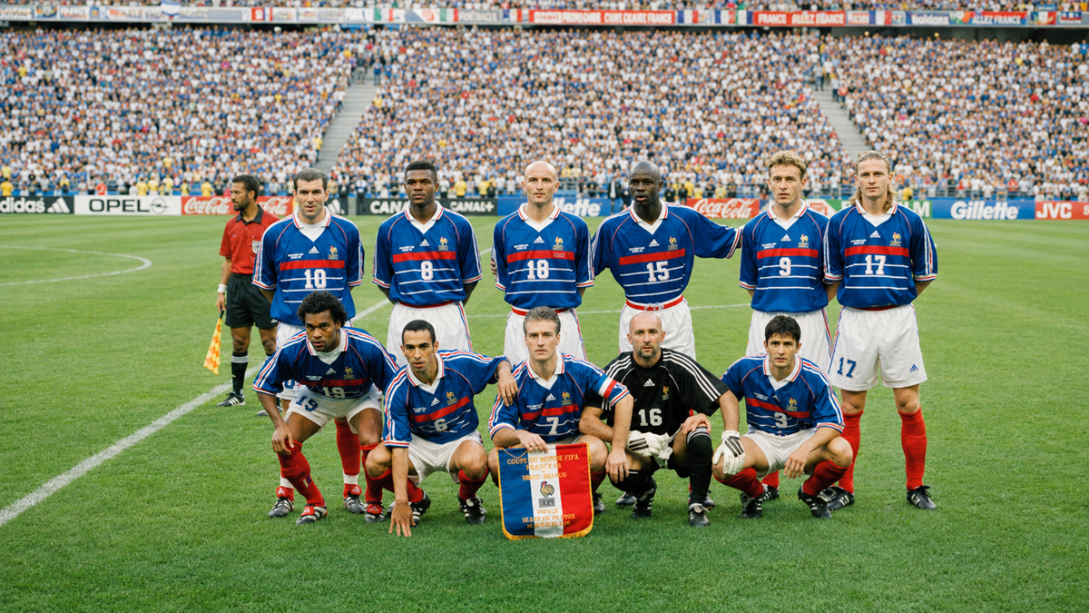 A soccer team in blue jerseys poses for a group photo on the field before a match, with a packed stadium and referee visible in the background. Eleven players are present, some standing and some kneeling.