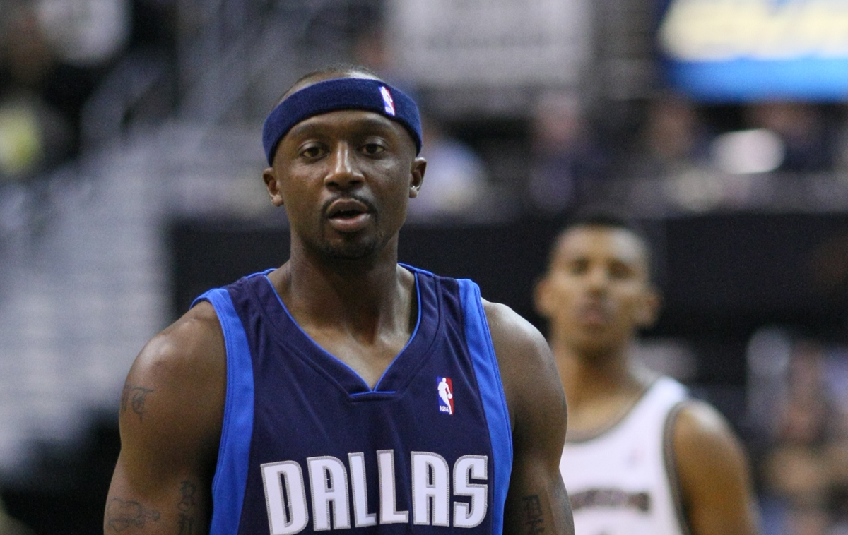 A basketball player in a blue Dallas jersey with a navy headband stands on the court, looking forward. The background is blurred with another player visible. Superstition.