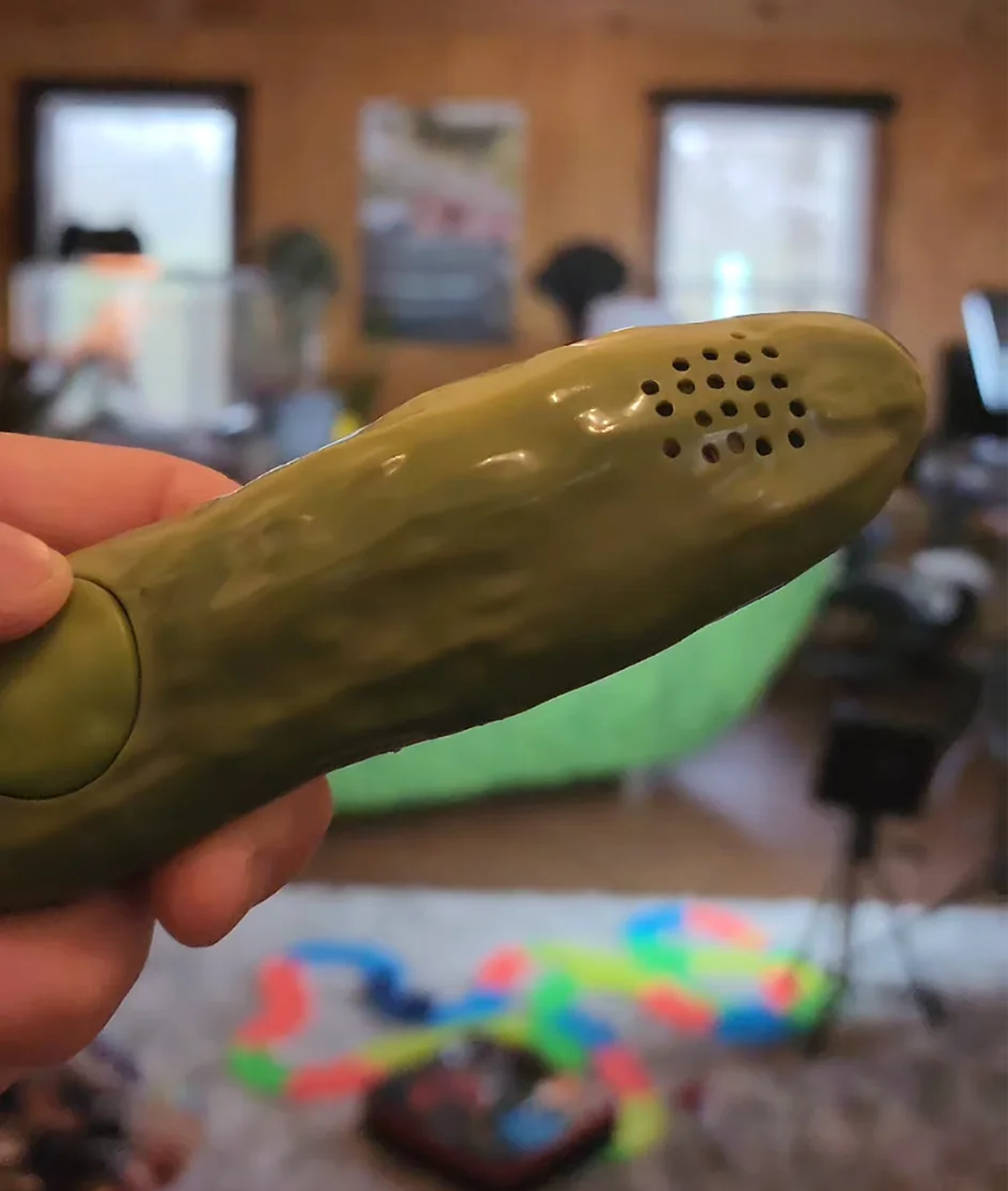 A hand holds a pickle-shaped object with speaker holes, resembling a toy or novelty item. The background is a blurred indoor room with various objects and furniture. Product