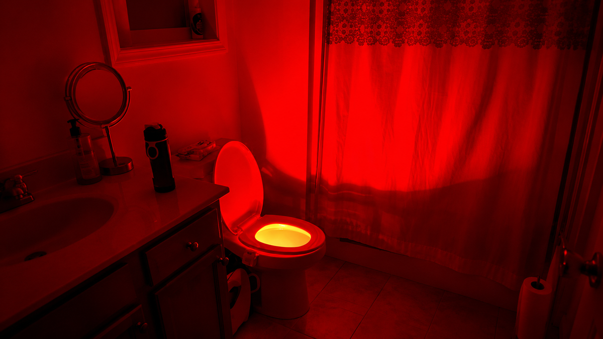 A bathroom lit with a red light; the toilet seat is open and glowing, casting red light around. The sink, mirror, and shower curtain are visible.