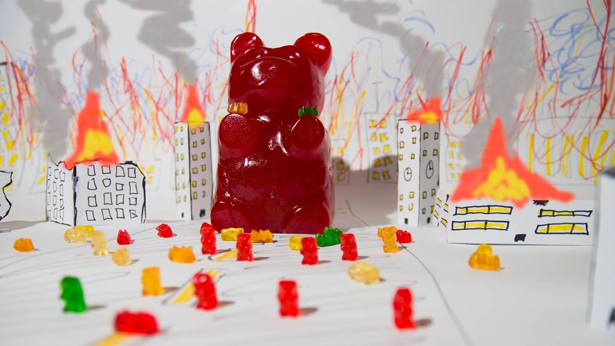A giant red gummy bear stands in the center of a city made from paper, surrounded by smaller gummy bears. The city is on fire, with hand-drawn flames and smoke on the paper buildings.