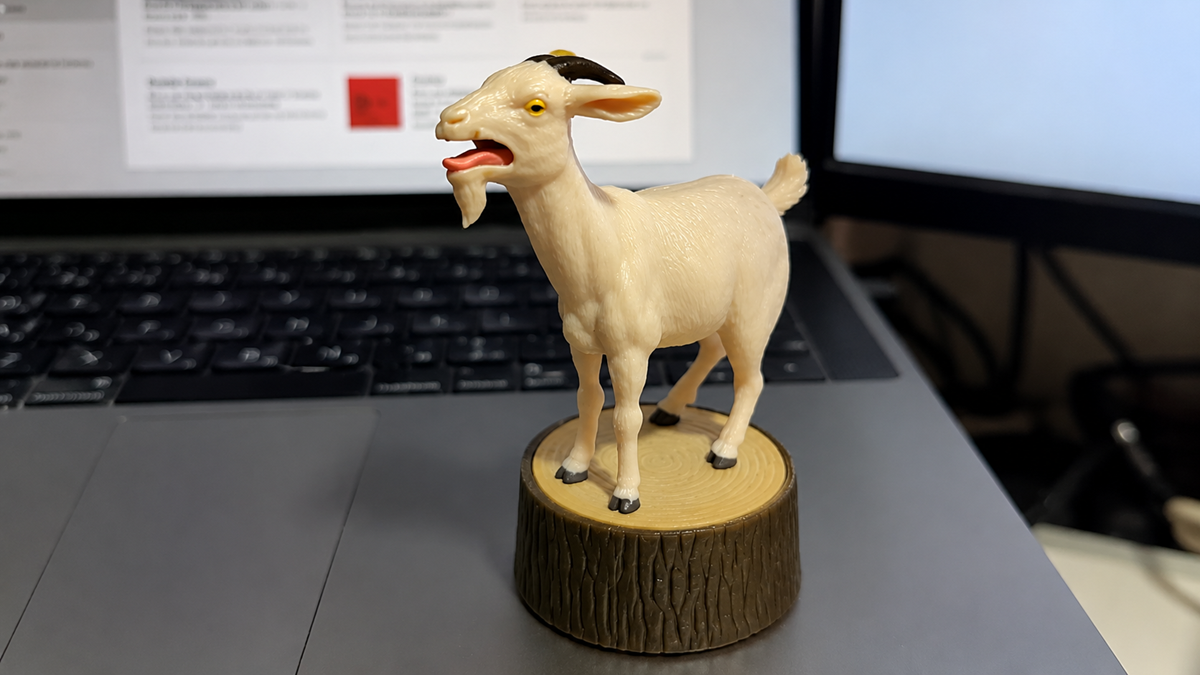 A small plastic goat figurine stands on a fake wooden stump in front of a laptop keyboard and monitor.
