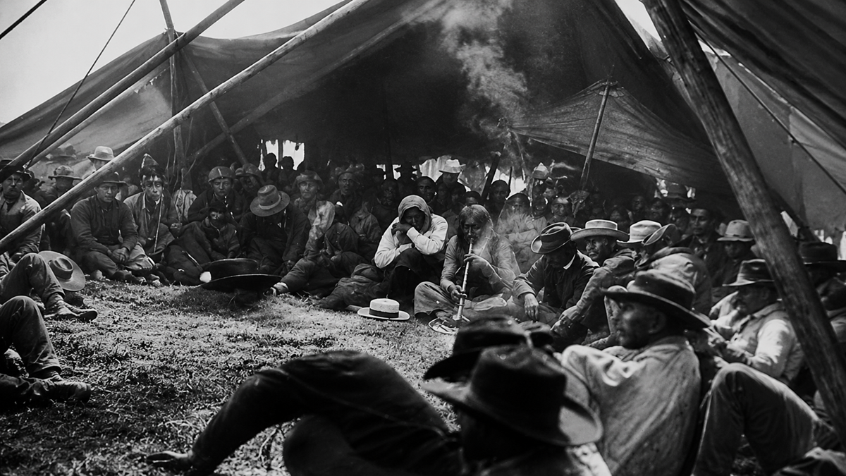A large group of men wearing hats sit closely together under a large canvas tent, with smoke rising in the center, creating a somber, contemplative atmosphere. The scene appears historical and outdoors.
