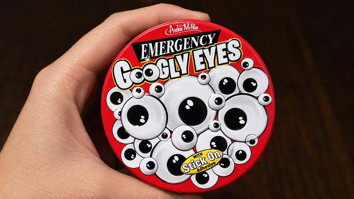 A hand holds a round red tin labeled "Emergency Googly Eyes" with cartoon googly eyes and text that reads "with Stick on Adhesive." The background is dark brown.