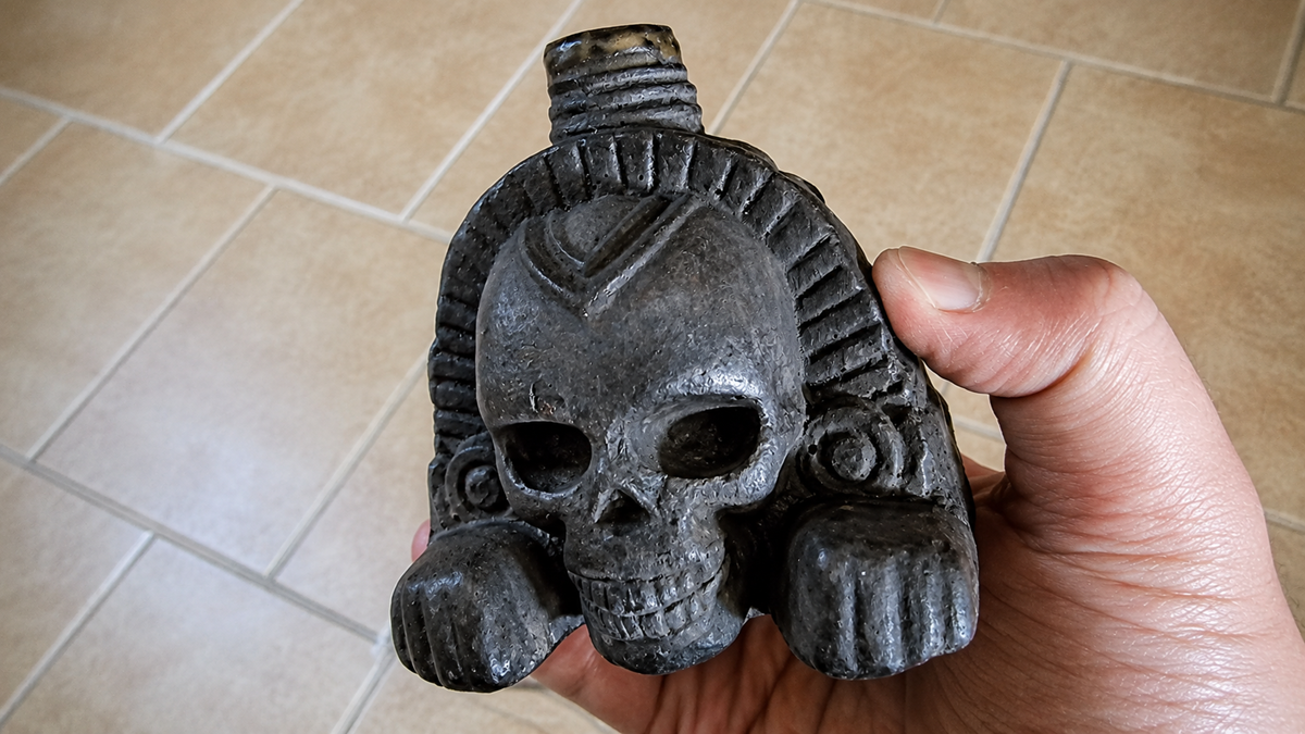 A hand holding a dark, carved stone object shaped like a skull with decorative details, possibly of cultural or historical significance, against a tiled floor background.