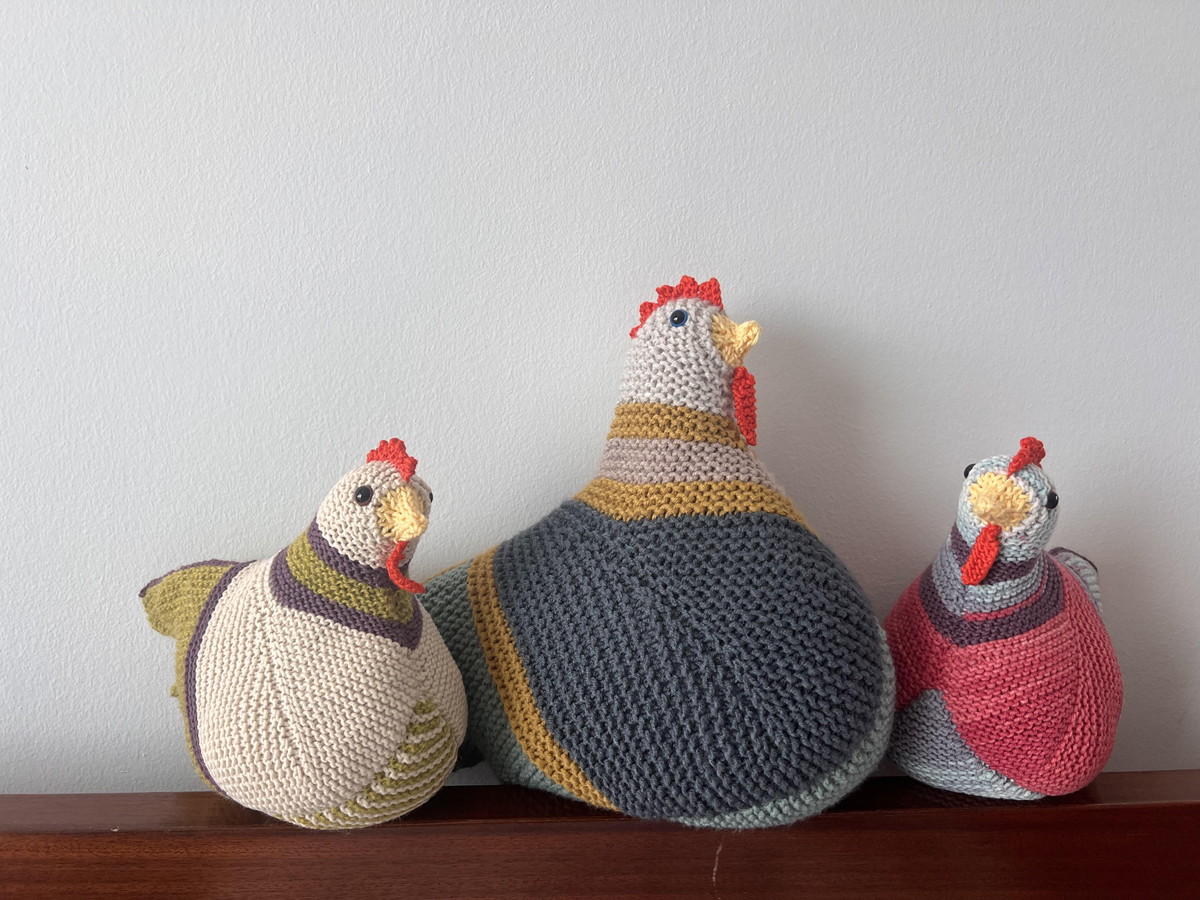 Three knitted chicken plushies with colorful patterns and red combs sit on a wooden surface against a light gray wall.