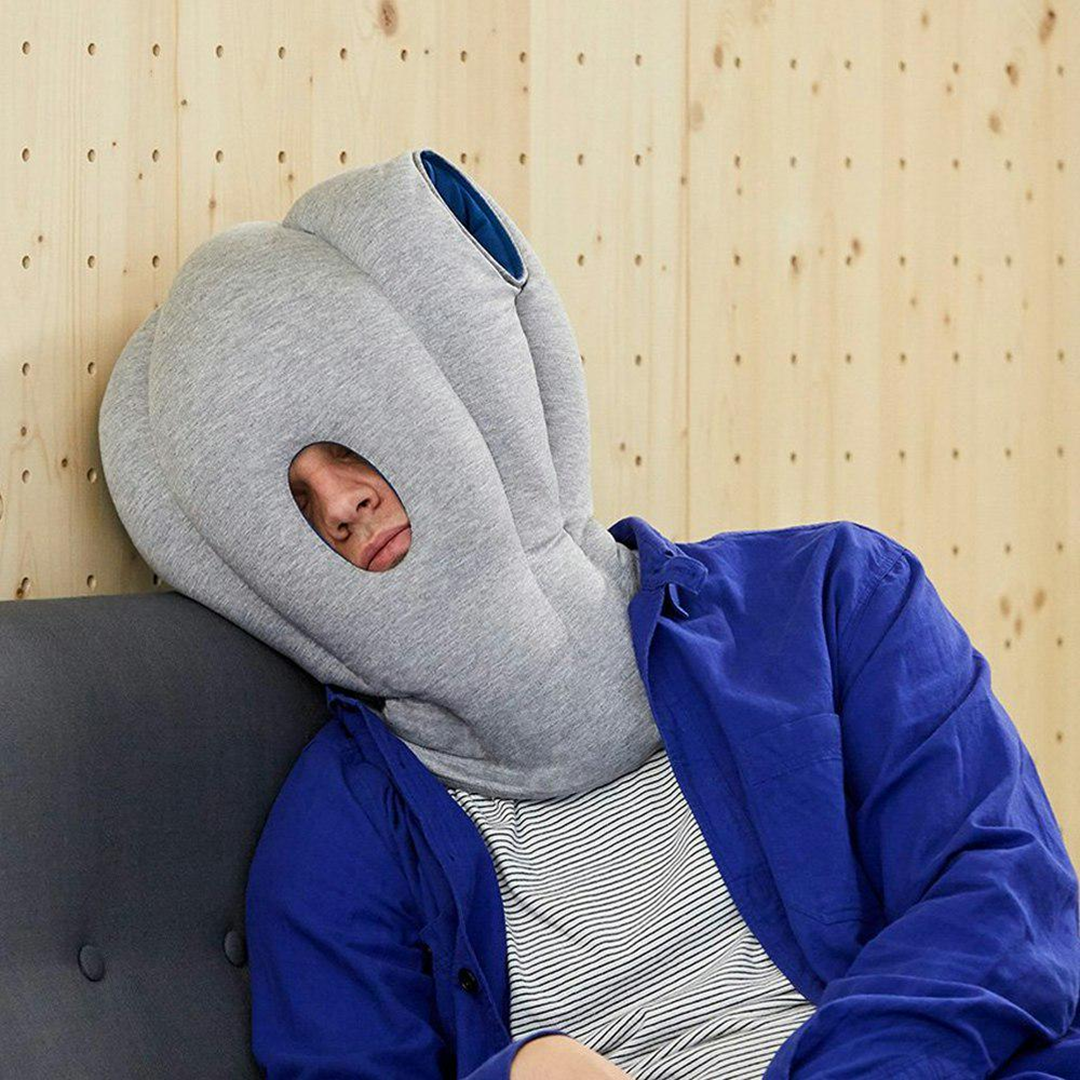 A person wearing a large, gray, padded hood with holes, resembling an octopus, is sitting on a couch and resting their head against a wall. They are dressed in a blue jacket and striped shirt.