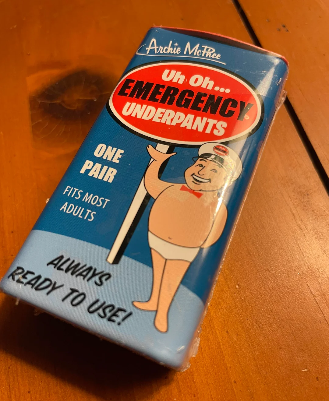 A package labeled "Emergency Underpants" with a cartoon man in underwear, text saying "One Pair, Fits Most Adults, Always Ready to Use!" lies on a wooden surface.