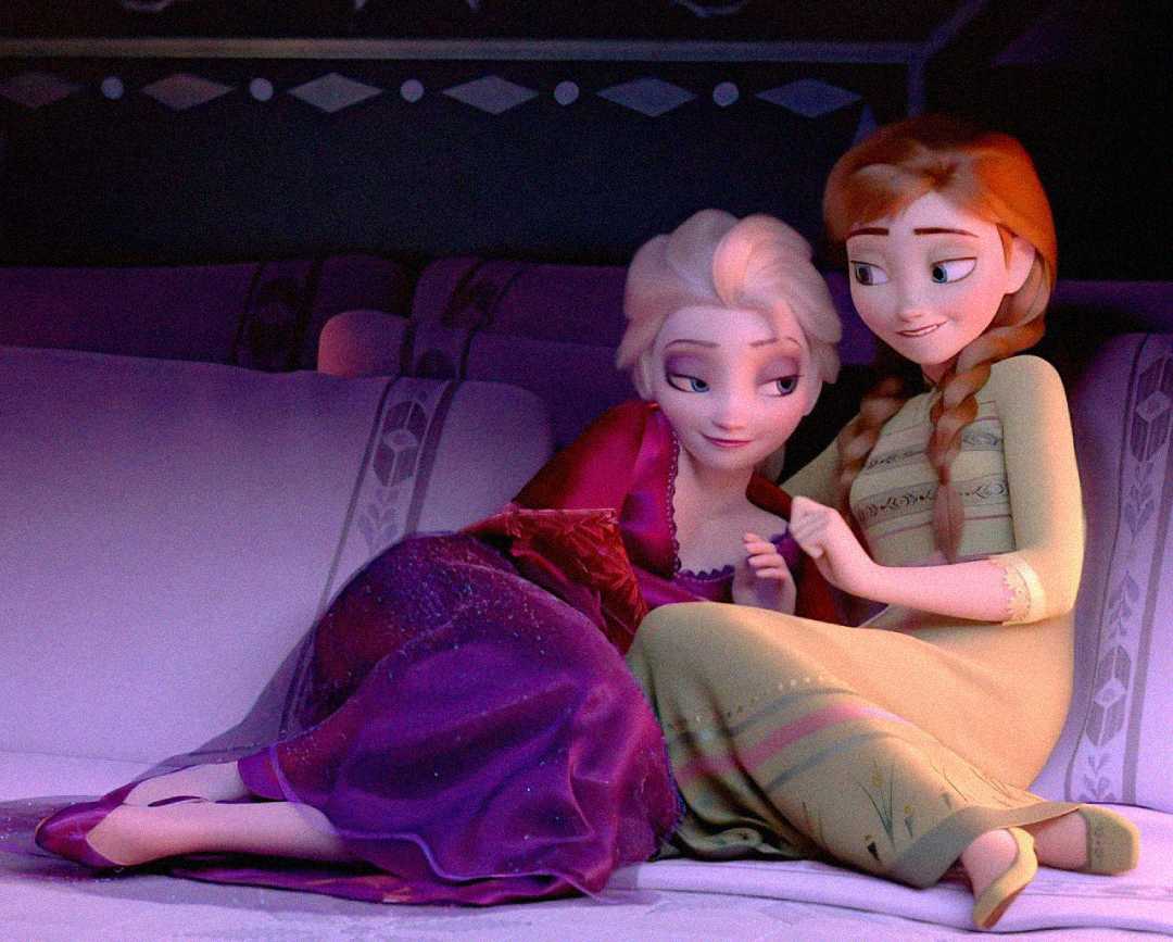 Elsa and Anna from Frozen sit close together on a couch, smiling warmly at each other. Elsa is wearing a purple dress and leaning on Anna, who is in a yellow dress with braids. The scene looks cozy and intimate.