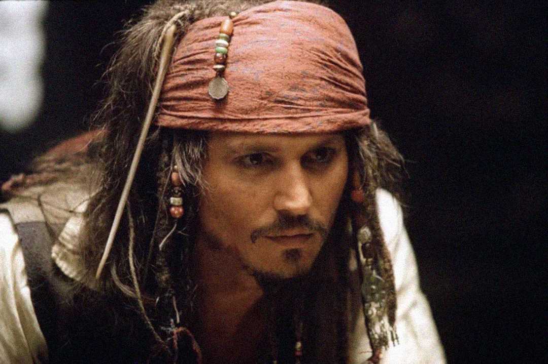 A man dressed as a pirate with a red bandana, beaded hair, and dark eyeliner looks intently forward. The background is dark and blurred, drawing focus to his serious expression and detailed costume.