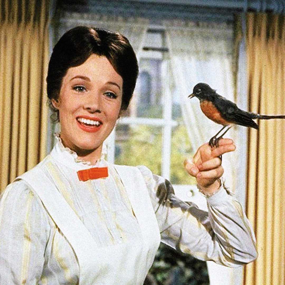 A smiling woman in a white dress and red bow tie stands indoors, holding her finger out as a small bird perches on it. Yellow curtains and a window with greenery outside are in the background.