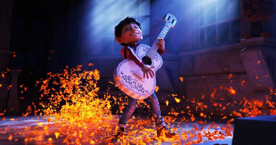 A young animated boy joyfully plays a white guitar as orange marigold petals swirl around him on a dimly lit stage, with a spotlight shining from above.