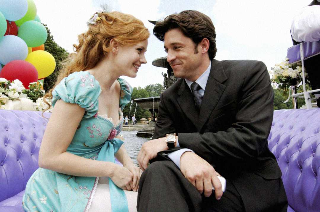A woman in a light blue dress and a man in a dark suit sit close together on a lavender couch outdoors, smiling at each other. Colorful balloons and a fountain are visible in the background.