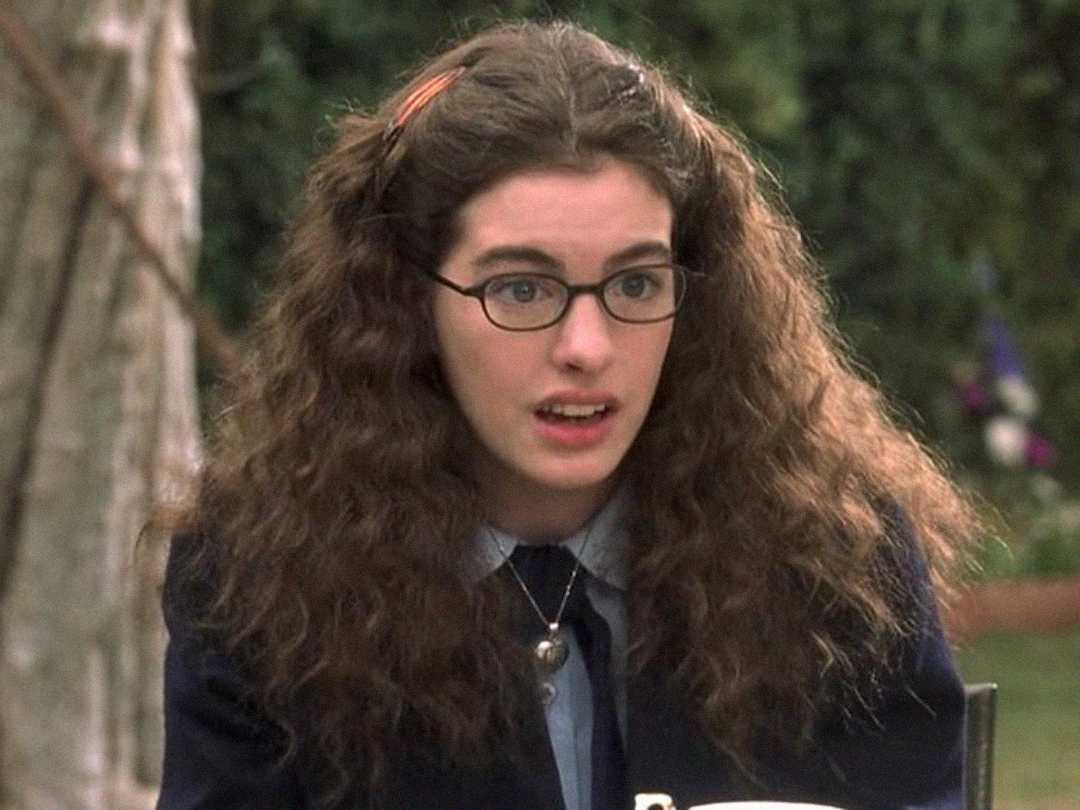 A young woman with long, curly brown hair, wearing glasses, a dark jacket, and a blue shirt sits outdoors, looking surprised or confused. There are trees and greenery in the blurred background.