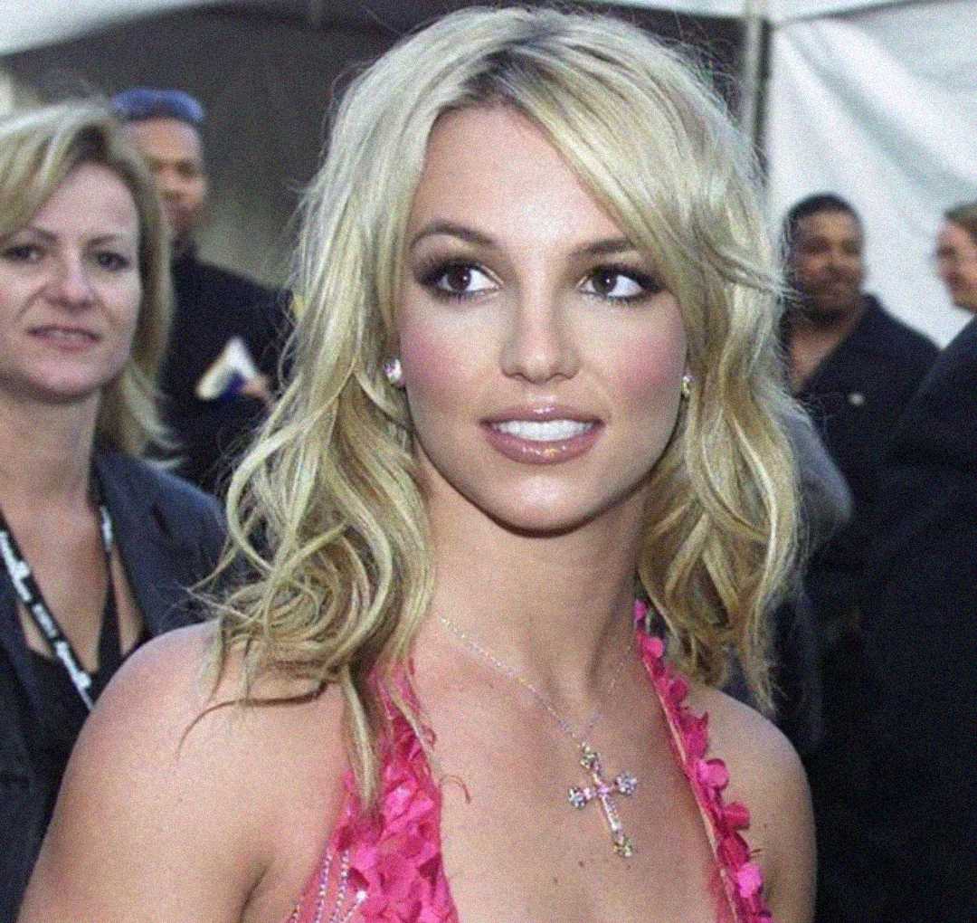 A woman with long blonde hair wearing a pink dress and a cross necklace looks slightly to the right, smiling softly, with people and a white tent blurred in the background.