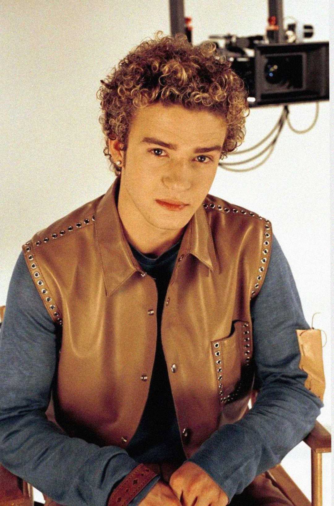 Young man with short, curly blond hair wearing a brown leather vest over a blue shirt sits on a chair in front of a beige background with some electronic equipment visible behind him.
