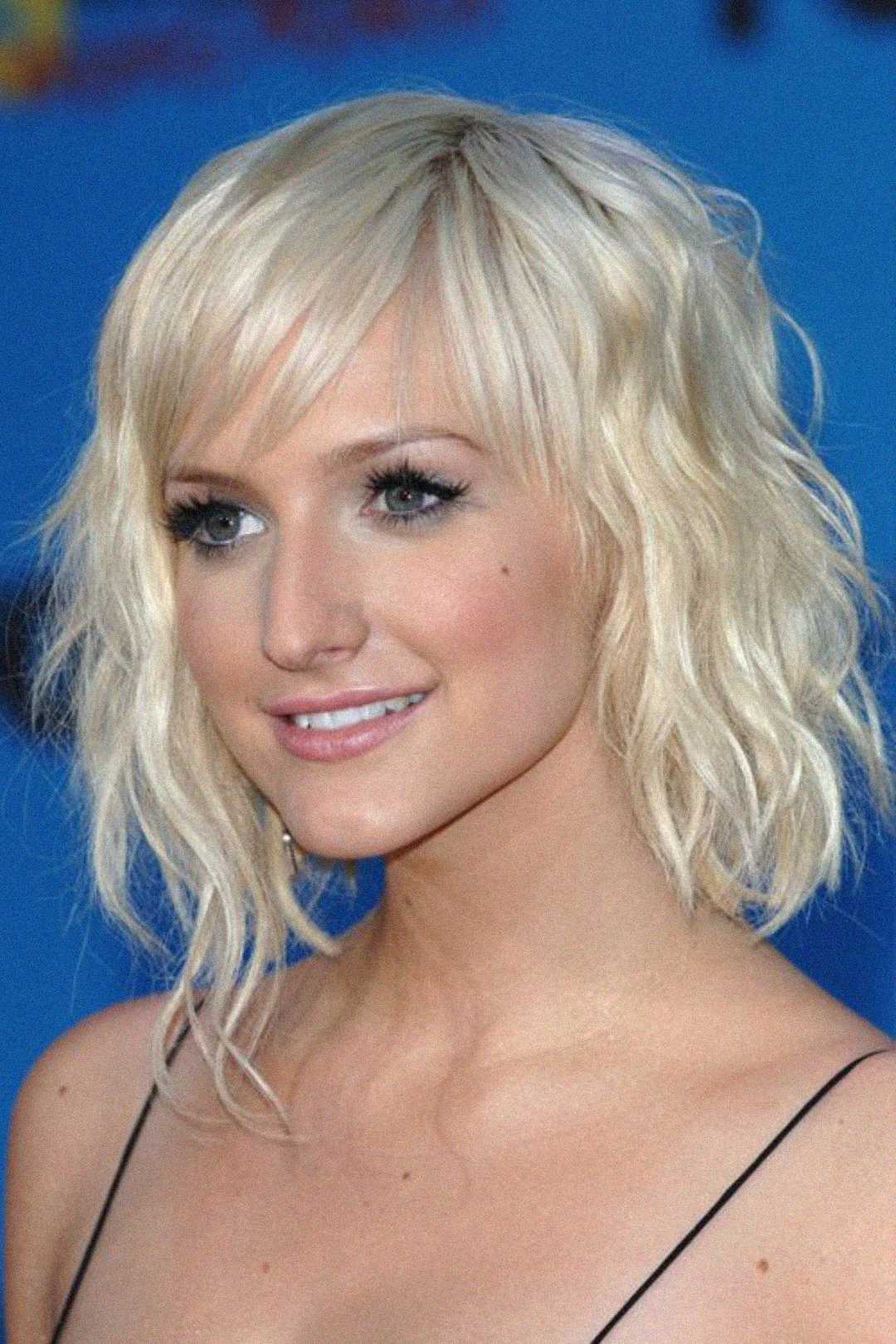 A woman with wavy, chin-length platinum blonde hair and side-swept bangs, wearing a black spaghetti strap top, poses in front of a blue background, smiling softly.