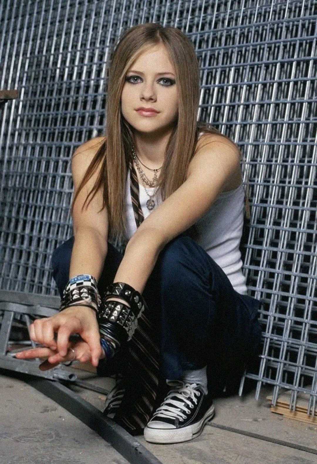 Young woman with straight brown hair, wearing a white tank top, dark pants, black Converse sneakers, and multiple bracelets, sits on the ground in front of a metal fence, looking at the camera with a neutral expression.