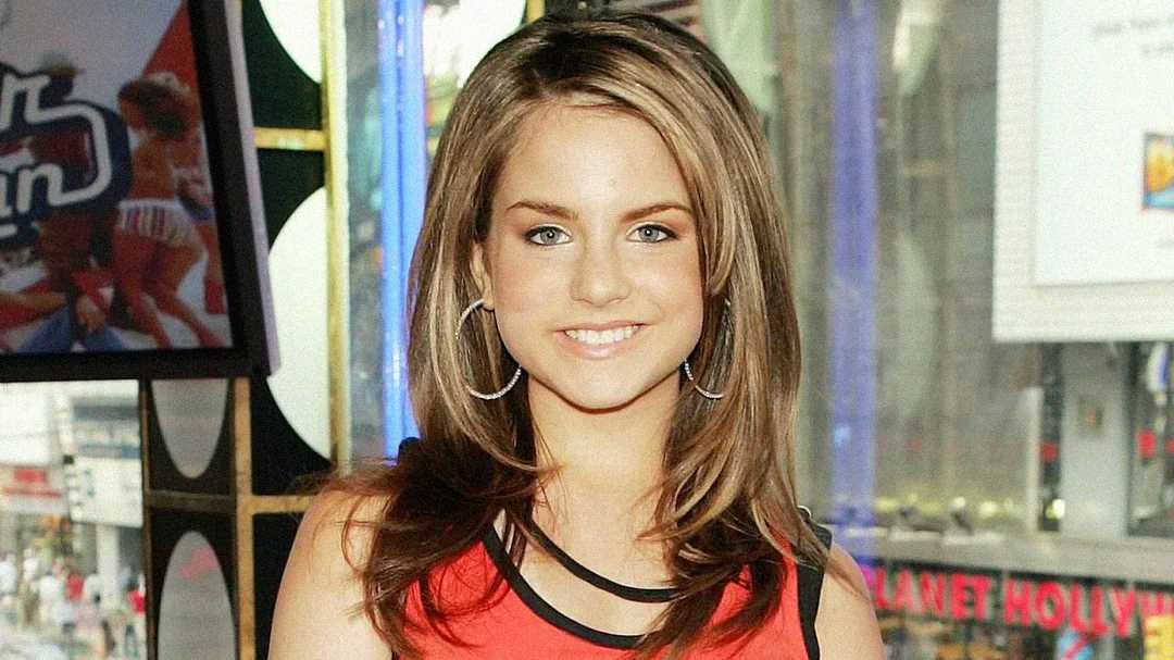 A young woman with straight, light brown hair and hoop earrings smiles at the camera while standing indoors in front of a window with city buildings visible outside.