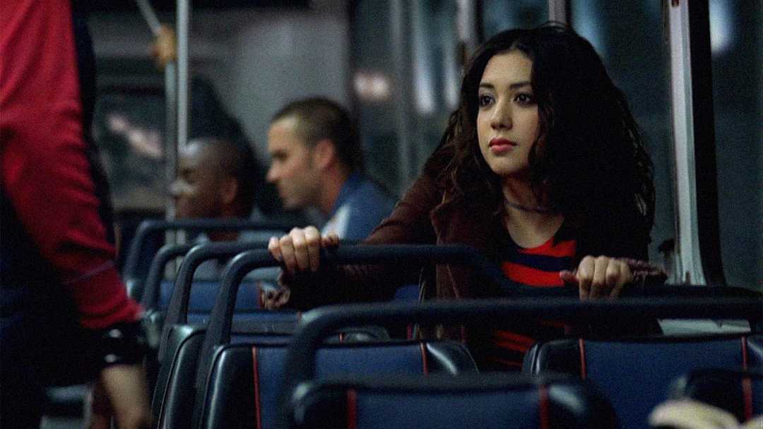 A young woman with long dark hair, wearing a red and black striped shirt, sits alone on a bus, holding the back of the seat in front of her and looking slightly to the side with a thoughtful expression.