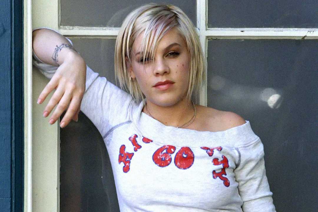 A person with short, platinum blonde hair leans casually against a window frame, wearing an off-shoulder white shirt with red text and a visible tattoo on their left wrist.