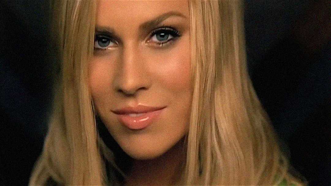 A close-up of a woman with long blonde hair, blue eyes, and glossy lips, looking directly at the camera with a slight smile. The background is dark and out of focus.